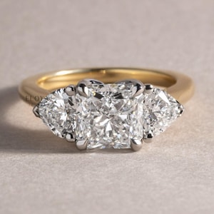 Peut inclure: Une bague en or avec trois diamants étincelants. Le diamant central est de forme carrée, flanqué de deux diamants triangulaires. Le sertissage est argenté et la monture est dorée. Le fond est neutre, mettant en valeur les pierres.