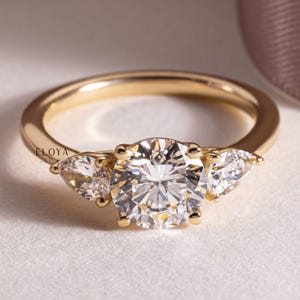 May include: A gold ring featuring a large, round, clear gemstone at the centre, with two smaller, pear-shaped gemstones either side. The ring is set on a white surface. The brand name "ELOYA JEWEL" is visible.