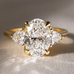 May include: A gold ring featuring a large, oval-cut diamond center stone, flanked by two pear-shaped diamonds. The ring has a classic, elegant design, perfect for engagement or special occasions.