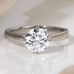 May include: A silver ring featuring a large, round, clear gemstone. The ring has a simple band and four prongs securing the gemstone. The brand name "ELOYA JEWEL" is visible on the ring.