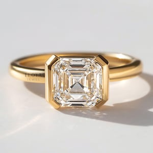 May include: A gold ring featuring a large, square-cut diamond. The diamond is set in a bezel setting, and the band is polished gold. The ring is displayed on a white surface. The text "ELOYA JEWEL" is visible on the ring.