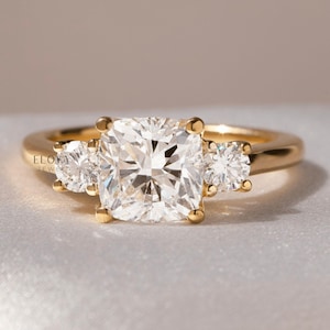 May include: A gold ring with a large, square-cut diamond center stone and two smaller side stones. The ring is set on a simple band and is displayed on a neutral background. The ring is from ELOYA JEWEL.