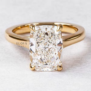 May include: A gold ring featuring a rectangular-cut diamond. The diamond has a brilliant, multifaceted surface. The gold band curves smoothly, with four prongs holding the diamond. The ring is set against a white background.