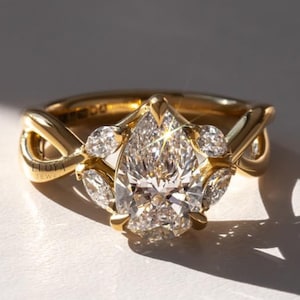 May include: A gold ring with a large, pear-shaped diamond center stone. The ring features smaller diamonds and an intricate band design. The ring is set on a white surface, with the brand name "ELOYA JEWEL" visible.