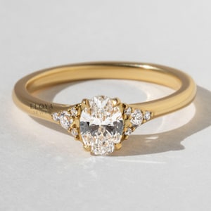 May include: A gold ring with a large oval-cut diamond center stone. The ring features smaller accent diamonds on either side of the center stone. The band is a simple, polished gold. The ring is set against a white background.