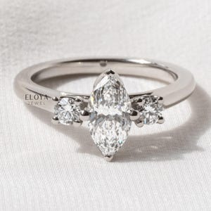 May include: A silver ring featuring a marquise-cut diamond center stone, flanked by two smaller round diamonds. The ring is set on a simple, polished band. The ring is displayed on a white surface.