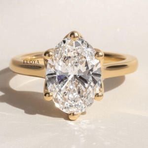 May include: A gold engagement ring featuring a large, oval-cut diamond. The diamond is secured by four gold prongs. The band is plain, polished gold. The ring is displayed against a soft, neutral background. The ring is a piece of jewellery.