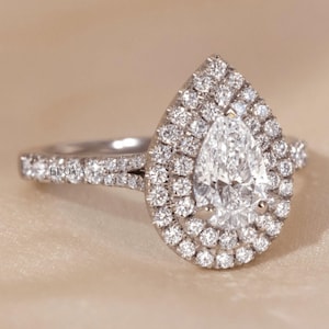 May include: A pear-shaped diamond ring with a double halo of smaller diamonds. The band is also encrusted with diamonds. The ring is set against a neutral background. The text "ELOYA JEWEL" is visible on the band.