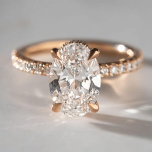 May include: A rose gold engagement ring with an oval-cut diamond center stone. The band is adorned with small, sparkling diamonds. The ring is set against a soft, neutral background. The brand name "ELOYA JEWEL" is visible on the band.