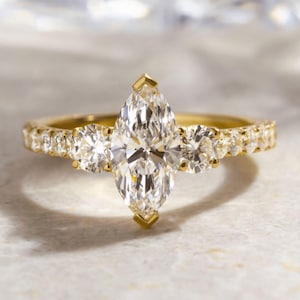 May include: A gold engagement ring with a marquise-cut diamond centre stone, accompanied by two round diamonds. The band is embellished with smaller diamonds. The ring is set against a neutral background, with the text "ELOYA JEWEL" visible.