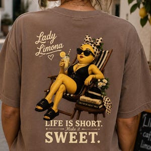 Op de afbeelding: Een taupekleurig t-shirt met een afbeelding van een geel personage in een zwart badpak, zonnebril en hoofddoek met stippen, zittend in een stoel. De tekst luidt Lady Limona, Life is Short. Make it Sweet, en Capri Dre Viing Not My Problem.