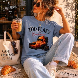 Op de afbeelding: Lichtblauw T-shirt met de tekst "TOO FLAKY for Commitment" en een afbeelding van een croissant met zonnebril. Een draagtas met "CAFÉ CROISSANT CHAOS" is zichtbaar. Een croissant en een kopje koffie zijn ook aanwezig.