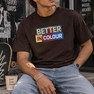 Op de afbeelding: Een donkerbruin t-shirt met korte mouwen en een grafische print met de tekst BETTER IN COLOUR in gele en witte letters tegen overlappende blauwe, gele en bordeauxrode rechthoekige blokken op het borstgedeelte van het kledingstuk.