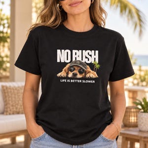 Op de afbeelding: Een zwart t-shirt met korte mouwen en een afbeelding van een golden retriever met een zonnebril en een baseballpet. Boven de hond staat de tekst NO RUSH en daaronder de tekst LIFE IS BETTER SLOWER, vergezeld door twee kleine groene palmbomen.