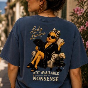 Op de afbeelding: Een blauw t-shirt met korte mouwen en een afbeelding op de achterkant van een figuur in een zwarte badjas, zonnebril en hoofddoek, zittend in een stoel met een waaier. De tekst luidt Lady Limona en NOT AVAILABLE for NONSENSE met hartjes.