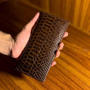 May include: A brown, textured wallet with a reptile skin pattern. The wallet is rectangular and held in a hand, showcasing its detailed design and colour. The wallet appears to be made of leather.