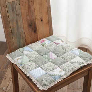 May include: A square patchwork chair cushion with a variety of patterned squares in green, white, and pastel colors. The cushion has a lace trim and is placed on a wooden chair.