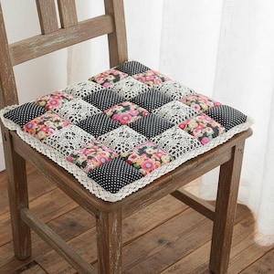 Puede incluir: Un cojín de silla cuadrado con un diseño de patchwork. El cojín presenta cuadrados alternos de lunares negros, estampados florales y encaje blanco. El cojín está bordeado con un ribete de encaje blanco y está sobre una silla de madera.
