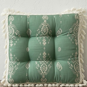 以下が含まれることがあります： 緑色の背景に白い花柄が施された正方形のフロアクッション。クッションはタフテッドデザインで、クリーム色の縁とタッセルでトリミングされています。この装飾的なクッションは屋内での使用に適しています。