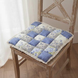 May include: A square chair cushion with a patchwork design. The cushion features alternating squares of blue polka dots, floral patterns, and white lace. The cushion is edged with a white lace trim and sits on a wooden chair.