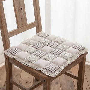 May include: A square chair cushion with a patchwork design. The cushion features alternating squares of beige fabric, brown and white gingham, and lace trim. The cushion is placed on a wooden chair.