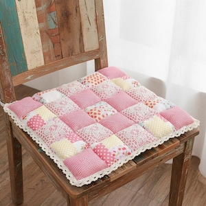 Puede incluir: Un cojín de silla cuadrado de patchwork con una variedad de cuadrados estampados en rosa y amarillo. El cojín tiene un ribete de encaje blanco y está colocado sobre una silla de madera. Los estampados incluyen lunares, cuadros y diseños florales.