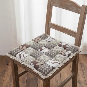 Puede incluir: Un cojín de silla cuadrado con un diseño de patchwork en tonos marrón, crema y blanco. El cojín presenta patrones florales y geométricos, con un ribete de encaje color crema. Está colocado sobre una silla de madera.