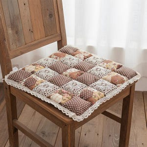 Könnte beinhalten: Ein quadratisches, gestepptes Stuhlkissen mit einem Patchwork-Design in Braun-, Creme- und floralen Farbtönen. Das Kissen hat eine weiße Spitzenborte und liegt auf einem Holzstuhl.