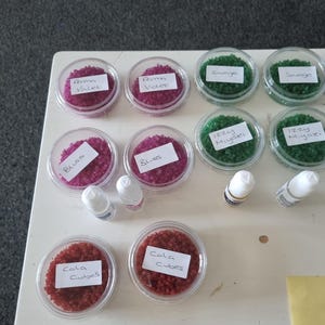 Könnte beinhalten: Draufsicht auf mehrere transparente Kunststoffbehälter, gefüllt mit farbigen Granulaten. Die Behälter sind mit Namen wie "Prima Violet", "Bliss", "Savage", "Izzy Miyaki" und "Cola Cubes" beschriftet. Kleine weiße Flaschen sind ebenfalls vorhanden.