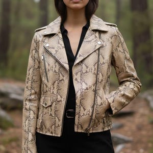 Könnte beinhalten: Beige und braune Lederjacke mit Schlangenmuster im klassischen Biker-Stil. Die Jacke hat einen Reverskragen, einen asymmetrischen Reißverschluss und mehrere Taschen. Die Person trägt ein schwarzes Hemd und eine Hose.