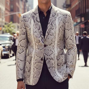 Może przedstawiać: Marynarka w wzór skóry węża z zapięciem na dwa guziki, noszona na czarnej koszuli. Kurtka ma neutralną paletę kolorów szarości, bieli i czerni. Zdjęcie zrobiono na ulicy miasta z ludźmi i samochodami w tle.