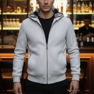 Puede incluir: Una sudadera con capucha blanca con cremallera y estampado de piel de serpiente, una capucha forrada de piel negra y una camisa negra debajo. La chaqueta tiene una cremallera frontal y dos bolsillos. El fondo es un bar con estanterías de botellas.