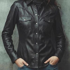 May include: Black leather shirt with two chest pockets and silver snap closures. The long-sleeved shirt has a classic collar and a fitted silhouette. The shirt is paired with blue jeans.