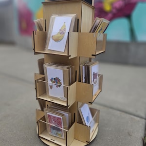 Puede incluir: Un expositor giratorio de madera lleno de tarjetas de felicitación. El expositor tiene varios estantes con divisores de plástico transparente. Las tarjetas presentan ilustraciones coloridas, incluyendo un plátano con gafas de sol y globos.