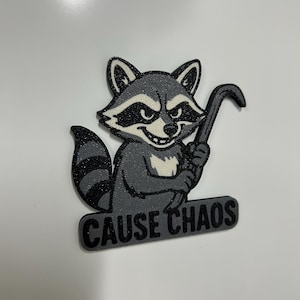May include: A cartoon raccoon in shades of grey and black, holding a crowbar. The text "CAUSE CHAOS" is below the raccoon. The image is likely a design for a sticker or patch, set against a white background.