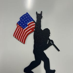 May include: Black metal silhouette of a Bigfoot-like figure holding a stick and an American flag. The flag features red and white stripes and a blue field with white stars. The figure is making a rock and roll hand gesture.