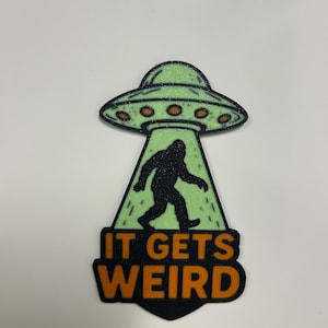 May include: A sticker featuring a green glitter UFO beaming up a black silhouette of Bigfoot. The text "IT GETS WEIRD" is in orange letters with a black outline, set against a black background.