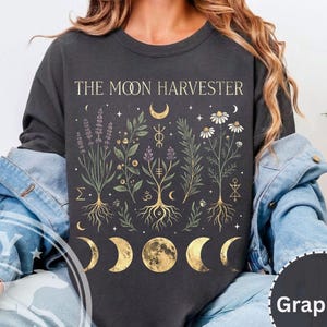 Może przedstawiać: Grafitowa koszulka z długim rękawem z napisem "THE MOON HARVESTER". Koszulka posiada botaniczny wzór z lawendą, stokrotkami i fazami księżyca w kolorze złotym. Koszulka jest w wygodnym, swobodnym stylu.
