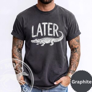 Pode incluir: Camiseta grafite com a palavra "LATER" e uma ilustra&ccedil;&atilde;o de um jacar&eacute; branco. A camisa tem mangas enroladas e um ajuste relaxado. Uma pe&ccedil;a de roupa confort&aacute;vel e casual.