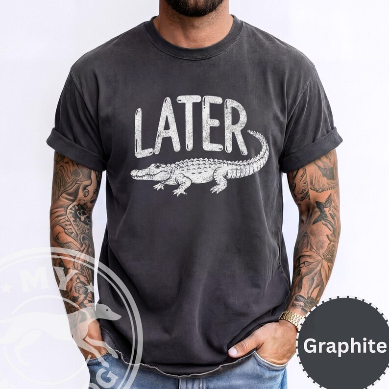 Pode incluir: Camiseta grafite com a palavra "LATER" e uma ilustra&ccedil;&atilde;o de um jacar&eacute; branco. A camisa tem mangas enroladas e um ajuste relaxado. Uma pe&ccedil;a de roupa confort&aacute;vel e casual.
