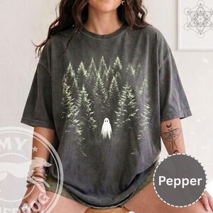 May include: A charcoal gray short-sleeve t-shirt featuring a graphic print of a dense forest of pine trees in shades of light green and white. A small, simple white ghost figure is positioned in the center of the forest graphic on the front.