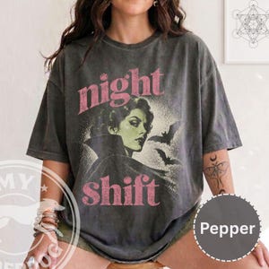 May include: A charcoal gray short-sleeve t-shirt featuring a graphic of a person with dark hair and a high-collared cape, accompanied by two flying bats. The text "night shift" is printed in pink, and a moth tattoo is visible on the arm.