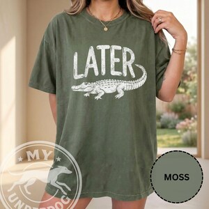 Pode incluir: Camiseta verde-oliva com a palavra "LATER" em letras brancas acima de uma ilustra&ccedil;&atilde;o de um jacar&eacute; branco. A camiseta tem um corte folgado e mangas curtas.