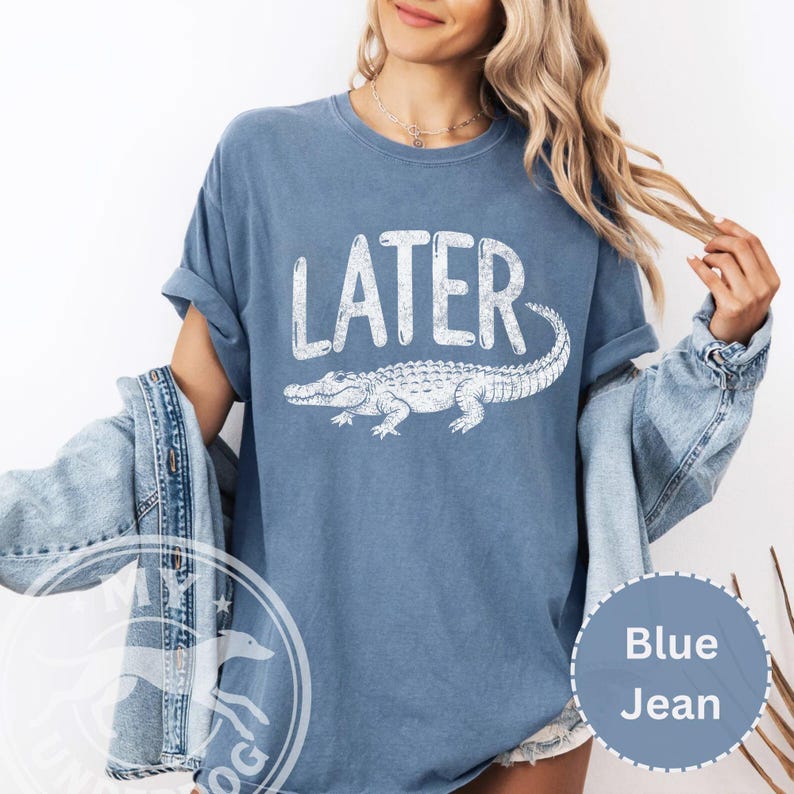 Pode incluir: Camiseta azul jeans com a palavra "LATER" em letras brancas sobre uma ilustra&ccedil;&atilde;o de um jacar&eacute; branco. A camiseta tem mangas curtas e um corte folgado.