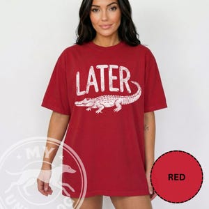 Pode incluir: T-shirt vermelha com a palavra "LATER" em branco, sobre uma ilustra&ccedil;&atilde;o de um jacar&eacute; branco. A t-shirt &eacute; de cor vermelha lisa e tem mangas curtas.