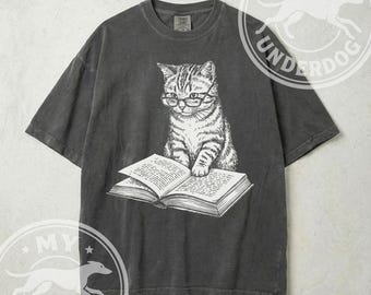 Könnte beinhalten: Dunkelgraues T-Shirt mit einer detaillierten Illustration einer Katze, die eine Brille trägt und ein Buch liest. Die Katze ist in sitzender Position dargestellt, mit ihren Pfoten auf dem offenen Buch. Das Shirt hat einen Vintage-Look.