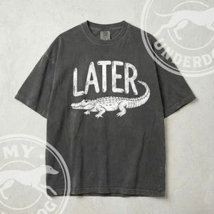 Pode incluir: T-shirt cinza escuro com a palavra "LATER" em branco acima de uma ilustra&ccedil;&atilde;o de um jacar&eacute; branco. A camisa tem mangas curtas e gola redonda.