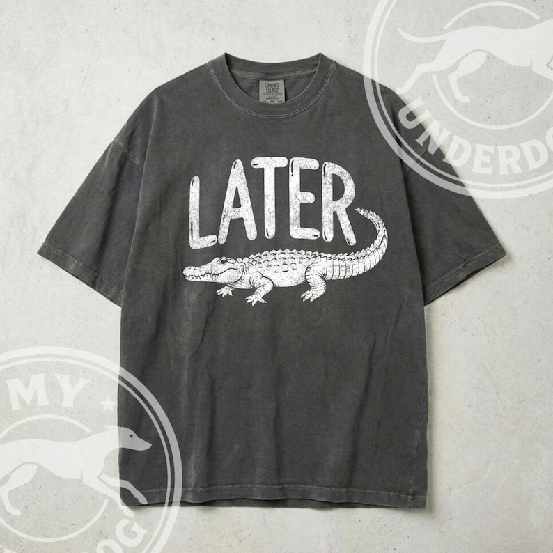 Pode incluir: T-shirt cinza escuro com a palavra "LATER" em branco acima de uma ilustra&ccedil;&atilde;o de um jacar&eacute; branco. A camisa tem mangas curtas e gola redonda.