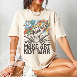 以下が含まれることがあります： クリーム色のTシャツ。デザインは、筆を持つ拳、鳩、花、虹が描かれています。拳の下には2丁の銃が交差しており、「MAKE ART NOT WAR」の文字が書かれています。