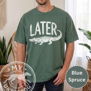 Pode incluir: Camiseta azul abeto com a palavra "LATER" acima de um gr&aacute;fico de jacar&eacute; branco. A camiseta de manga curta e gola redonda tem uma cor s&oacute;lida com um gr&aacute;fico desgastado. Estilo casual.